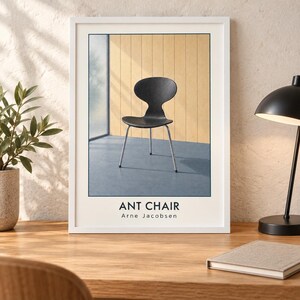 May include: A framed wall art print featuring a black Ant Chair by Arne Jacobsen. The chair has a curved seat and backrest with thin metal legs, set against a light wood paneled wall and a blue floor. The text reads ANT CHAIR Arne Jacobsen.