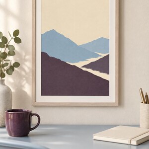 May include: A framed wall art print featuring a layered mountain landscape illustration. The artwork displays a pale cream sky above light blue mountain peaks, with dark purple foreground slopes and a winding white path through the center.