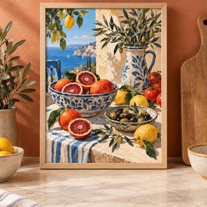 Op de afbeelding: Een ingelijste kunstdruk met een mediterrane sc&egrave;ne met een blauw-wit patroon kom gevuld met bloedsinaasappels, een bijpassende kan met olijftakken, een kom met olijven, citroenen en tomaten op een tafel met uitzicht op de zee.