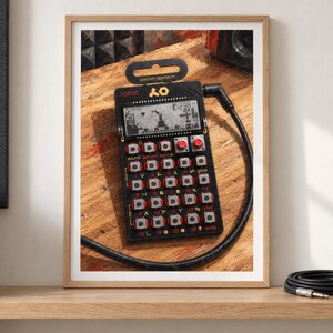 May include: A black Teenage Engineering PO-28 Robot pocket operator synthesiser featuring a monochrome LCD screen, a grid of twenty-three silver buttons with red accents, and a black audio cable plugged into the top right side of the device.