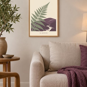 May include: A rectangular wall art print in a light wood frame featuring a stylised green fern leaf overlapping a dark purple mountain landscape with a winding white river. The word LAOIS is printed in black capital letters at the bottom centre.