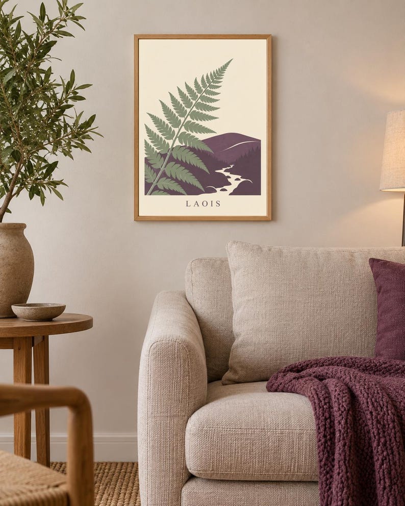 May include: A rectangular wall art print in a light wood frame featuring a stylised green fern leaf overlapping a dark purple mountain landscape with a winding white river. The word LAOIS is printed in black capital letters at the bottom centre.
