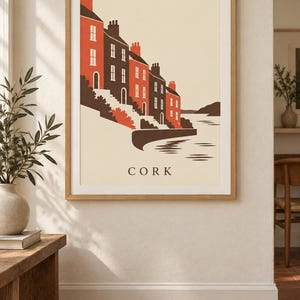 Op de afbeelding: Een ingelijste kunstdruk met een gestileerde illustratie van gebouwen langs een waterkant in Cork. Het kunstwerk gebruikt een palet van cr&egrave;me, donkerbruin en oranje, met een rij huizen met schoorstenen, trappen en water. De tekst CORK staat gecentreerd onderaan.