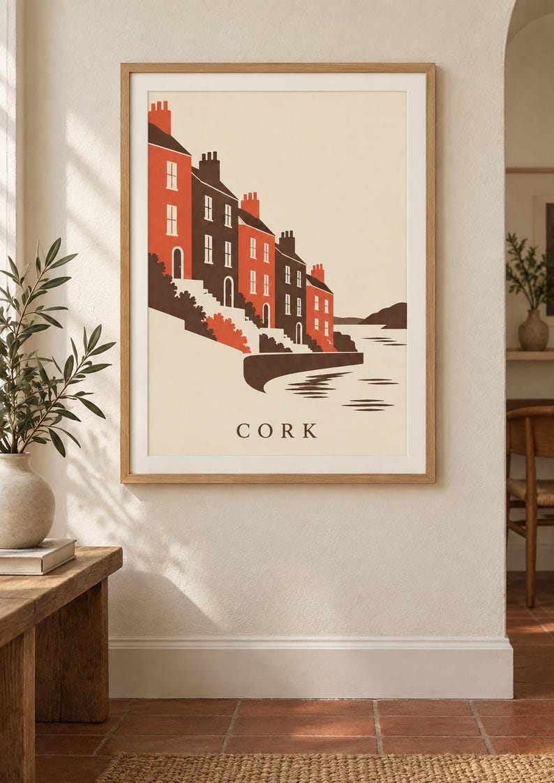 Op de afbeelding: Een ingelijste kunstdruk met een gestileerde illustratie van gebouwen langs een waterkant in Cork. Het kunstwerk gebruikt een palet van cr&egrave;me, donkerbruin en oranje, met een rij huizen met schoorstenen, trappen en water. De tekst CORK staat gecentreerd onderaan.