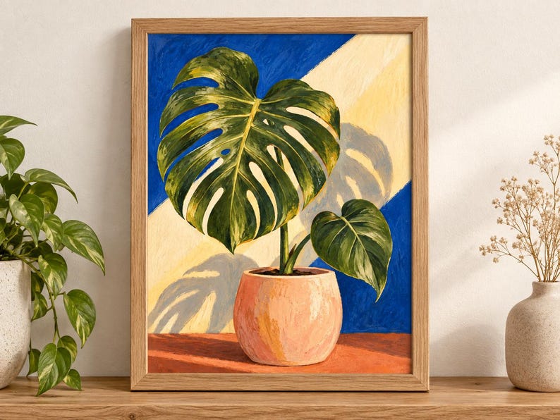 Puede incluir: Un cuadro enmarcado de una planta monstera verde en una maceta color melocot&oacute;n. El fondo presenta una divisi&oacute;n diagonal de azul intenso y amarillo p&aacute;lido, creando una sombra de las grandes hojas divididas sobre la secci&oacute;n amarilla de la pared.