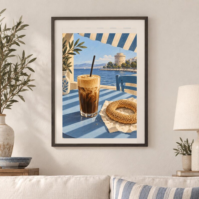 May include: A framed wall art print depicting a glass of iced coffee with a black straw and a sesame seed bagel on a blue table. The background shows a coastal scene with a white tower, blue sea, and a striped awning under a bright sky.
