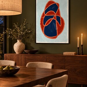 May include: A framed wall art print featuring an abstract composition with a large, irregular red and dark blue intertwined shape centered on a light blue background. The artwork is enclosed in a thin black frame and displayed on a wall.