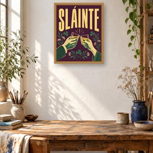 Pu&ograve; includere: Una stampa artistica da parete rettangolare con sfondo viola scuro e la parola SL&Aacute;INTE in grandi lettere gialle. L'illustrazione raffigura due mani che brindano con bicchieri pieni di un liquido dorato, circondate da motivi di trifogli e fiori verdi.