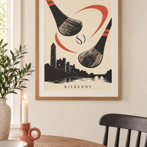 Peut inclure: Une impression d'art encadr&eacute;e pr&eacute;sentant une illustration stylis&eacute;e de deux crosses de hurling noires et d'une balle en mouvement au-dessus d'une silhouette du ch&acirc;teau et de la tour ronde de Kilkenny. Fond cr&egrave;me avec des accents rouges et le texte KILKENNY.
