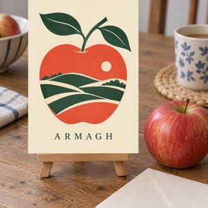 May include: A rectangular print on a small wooden easel featuring a graphic illustration of a red apple with two green leaves. Inside the apple shape is a landscape of green rolling hills and a white sun. The word ARMAGH is printed in green below.
