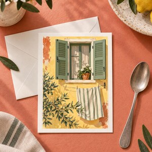 May include: A rectangular greeting card featuring an illustration of a yellow wall with an open window, green shutters, a potted plant, and a striped towel hanging on a clothesline, accompanied by a white envelope, a silver spoon, and lemons.