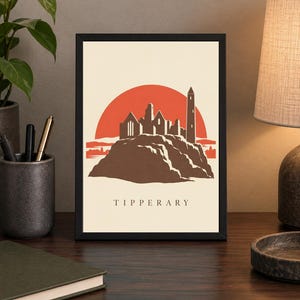 May include: A framed art print featuring a stylized illustration of the Rock of Cashel in Tipperary, Ireland. The design shows a brown stone castle silhouette against a large orange sun on a cream background, with the word TIPPERARY printed below.