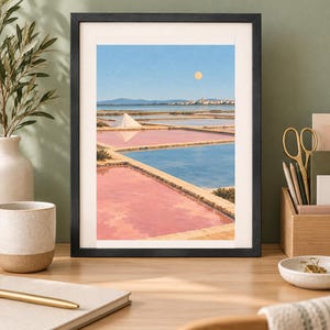 May include: A framed art print depicting a landscape of salt pans with rectangular pools of pink and blue water, a white salt mound, a distant town, and a pale yellow sun in a clear blue sky, all enclosed in a simple black frame.