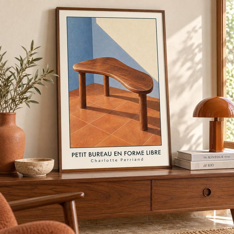 May include: A framed art print featuring a wooden table with a free-form top and three cylindrical legs. The background shows a blue and cream wall above a terracotta tiled floor. The text PETIT BUREAU EN FORME LIBRE Charlotte Perriand is printed below.