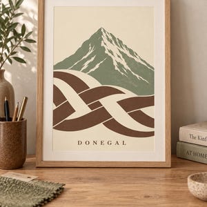 K&ouml;nnte beinhalten: Ein gerahmter Kunstdruck mit einem stilisierten gr&uuml;nen Berg mit wei&szlig;en Schneeakzenten &uuml;ber einem braun-cremefarbenen, ineinander verschlungenen Knotenmuster. Das Wort DONEGAL ist in schwarzen Gro&szlig;buchstaben unten mittig auf dem cremefarbenen Hintergrund gedruckt.