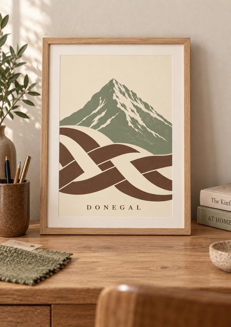 K&ouml;nnte beinhalten: Ein gerahmter Kunstdruck mit einem stilisierten gr&uuml;nen Berg mit wei&szlig;en Schneeakzenten &uuml;ber einem braun-cremefarbenen, ineinander verschlungenen Knotenmuster. Das Wort DONEGAL ist in schwarzen Gro&szlig;buchstaben unten mittig auf dem cremefarbenen Hintergrund gedruckt.