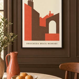 K&ouml;nnte beinhalten: Ein gerahmter Kunstdruck mit dem Titel DROGHEDA BRICK MEMORY, der eine geometrische Illustration von Geb&auml;uden in Dunkelbraun, gebranntem Orange und Creme zeigt. Das Bild zeigt stilisierte D&auml;cher, Schornsteine und einen Torbogen.