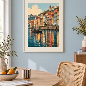 May include: A framed wall art print depicting a coastal town with colorful buildings along a waterfront, a small boat floating on the water, and reflections of the structures on the surface, all rendered in a painterly style with warm tones.