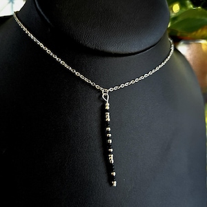 Può includere: Una delicata collana con catena d'argento e pendente verticale. Il pendente presenta una serie di piccole perline nere intervallate da perline color argento. La collana è esposta su un busto di gioielli nero.