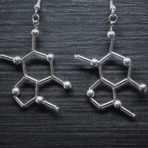 Peut inclure: Une paire de boucles d'oreilles en argent en forme de molécule de caféine. Les boucles d'oreilles sont faites de petites perles d'argent reliées par un fil d'argent fin.