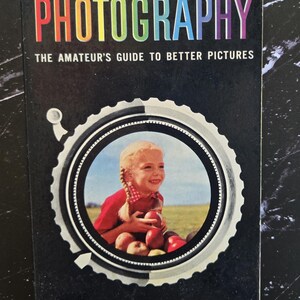 May include: Vintage book cover for "Photography: The Amateur's Guide to Better Pictures." The title is in rainbow colors. The cover features a girl holding apples inside a camera lens illustration. "A Golden Handbook" is printed at the bottom.