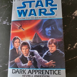 May include: A book cover for "Star Wars: Dark Apprentice", volume 2 of the Jedi Academy Trilogy. The cover features a dark blue and red background with a scene of Luke Skywalker, Han Solo, and Princess Leia. The text "The Blockbuster Saga Continues" is at the top of the cover, and the title "Dark Apprentice" is at the bottom.