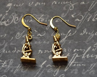 May include: A pair of gold-toned earrings featuring a miniature microscope charm. The earrings have a simple hook design.