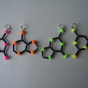 Könnte beinhalten: Vier verschiedene farbige Perlenketten, jede mit einer schwarzen Schnur und einer silbernen Schlaufe. Die Ketten sind sechseckig mit einem kleinen Kreis in der Mitte. Die Farben der Perlen sind rosa, orange, grün und lindgrün.