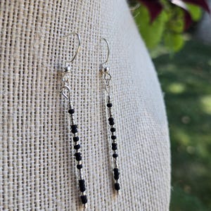 May include: A pair of dangle earrings with silver-colored hooks and beaded strands. The strands alternate small black beads with silver-colored beads, creating a vertical striped pattern. The earrings are displayed on a beige fabric.