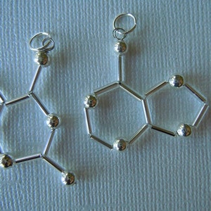 Könnte beinhalten: Zwei silberne Halsketten mit Anhänger in Form von Molekülstrukturen. Die Anhänger bestehen aus kleinen silbernen Perlen, die durch silberne Stäbe verbunden sind.