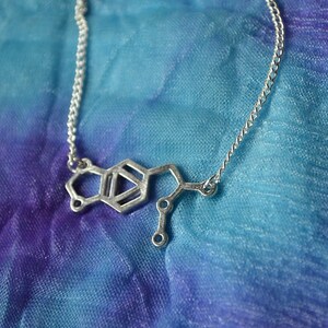 May include: A silver necklace with a pendant shaped like the chemical structure of dopamine. The necklace is on a blue and purple fabric background.