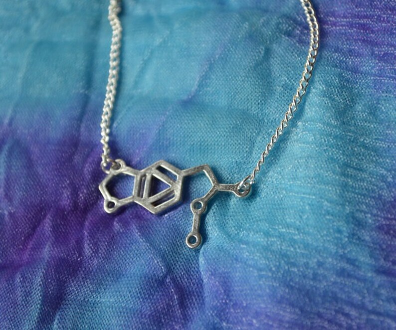 May include: A silver necklace with a pendant shaped like the chemical structure of dopamine. The necklace is on a blue and purple fabric background.