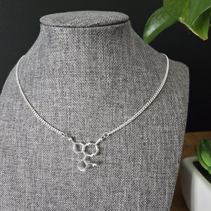 May include: A silver chain necklace with a silver pendant shaped like a chemical structure. The necklace is displayed on a gray mannequin.