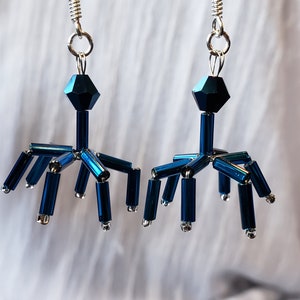 Puede incluir: Un par de pendientes abstractos en azul oscuro. Cada pendiente tiene una cuenta facetada, una varilla vertical central y varias varillas horizontales con tapas plateadas. Los pendientes cuelgan de ganchos plateados, creando un dise&ntilde;o sim&eacute;trico. Miden aproximadamente 5 cm de largo.