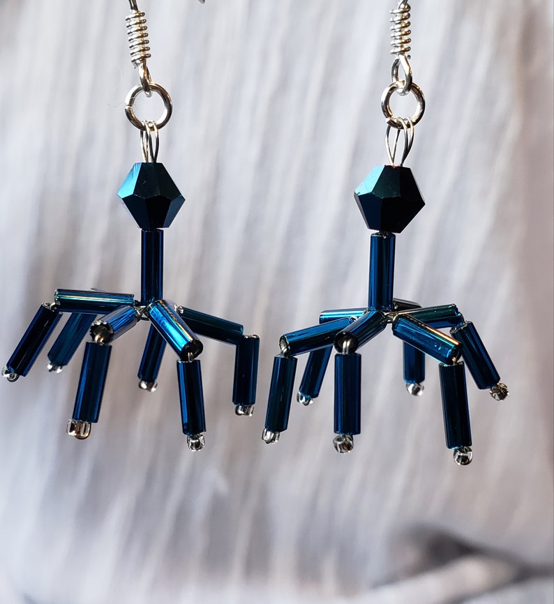 Puede incluir: Un par de pendientes abstractos en azul oscuro. Cada pendiente tiene una cuenta facetada, una varilla vertical central y varias varillas horizontales con tapas plateadas. Los pendientes cuelgan de ganchos plateados, creando un dise&ntilde;o sim&eacute;trico. Miden aproximadamente 5 cm de largo.
