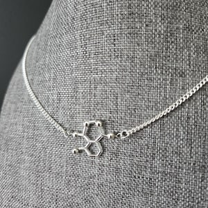 May include: A silver necklace with a pendant shaped like the chemical structure of serotonin. The necklace is on a grey fabric mannequin.