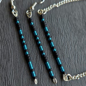 May include: A handmade jewellery set comprising earrings and a necklace. The earrings and necklace feature a beaded design with black and teal blue beads. The earrings have silver-coloured ear wires, and the necklace has a silver-coloured chain.