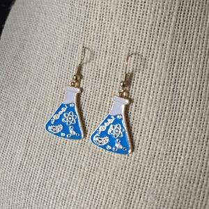 Può includere: Orecchini color oro con un design a forma di beuta in smalto blu. Le beute hanno una parte superiore bianca e sono decorate con grafiche a tema scientifico. Gli orecchini sono esposti su una superficie strutturata di colore neutro.