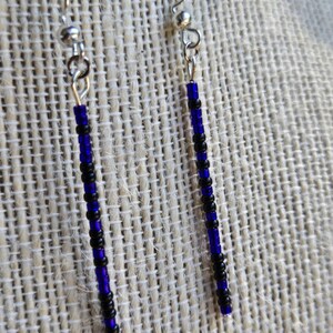 May include: Dangle earrings with a beaded design. The earrings feature a combination of black and blue beads strung together, creating a vertical striped pattern. The ear wires are silver-colored.