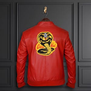 Pode incluir: Um casaco vermelho brilhante com um grande emblema circular nas costas. O emblema apresenta uma cobra preta com contorno amarelo e o texto Cobra Kai em letras vermelhas. O casaco tem uma gola subida e punhos e bainha canelados.