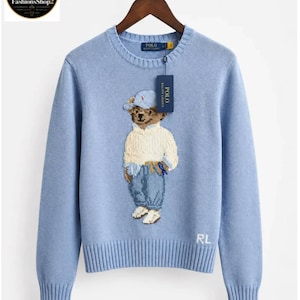 May include: A light blue long-sleeve crewneck sweater featuring an embroidered bear wearing a cap, a cream-colored sweater, and blue jeans. The sweater has a Polo Ralph Lauren label at the neck, a hanging brand tag, and the letters RL embroidered in white.