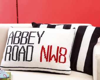 May include: Two decorative throw pillows on a white sofa. One pillow is white with black text that reads "ABBEY ROAD NW8" in black and red. The other pillow has black and white stripes. The background is a red wall.