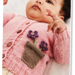 以下が含まれることがあります： ピンクのニットのベビーカーディガン。前面に茶色のニットの花瓶と紫の花が飾られています。