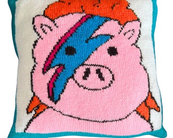 May include: A pink knitted pillow with a black outline of a pig's face. The pig has a blue, red, and pink lightning bolt design across its face. The pillow has a turquoise border.