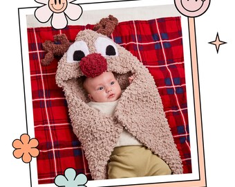 May include: A baby wearing a brown knitted reindeer hooded blanket on a red and black plaid blanket.