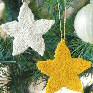 Pode incluir: Duas decorações de estrela de malha penduradas em uma árvore de Natal. Uma estrela é creme, a outra é amarela. Ambas as estrelas têm cordas douradas para pendurar. O fundo inclui agulhas de pinheiro verdes e uma bola branca.