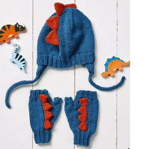 May include: A blue knitted dinosaur-themed hat with ear flaps and matching fingerless gloves. The hat and gloves feature orange dinosaur spikes. Wooden dinosaur toys are scattered around the set.