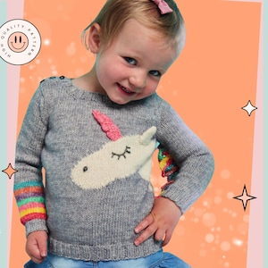 Può includere: Un maglione di lana grigio con un'applicazione di unicorno bianco e strisce arcobaleno sulle maniche. Il maglione è per un bambino e presenta il testo "Unicorno e arcobaleno".