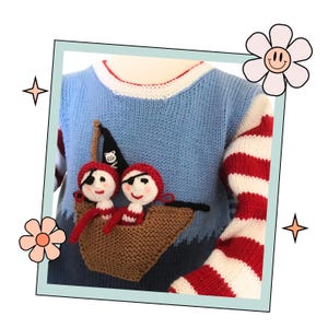 Könnte beinhalten: Ein blauer Strickpullover mit rot-weiß gestreiften Ärmeln. Der Pullover zeigt ein braunes Strickboot mit zwei rot-weißen Strickpiraten an Bord.