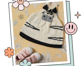 May include: A white knitted baby dress with black stripes and a zebra face design. The dress has a black trim and is laying on a floral patterned surface.